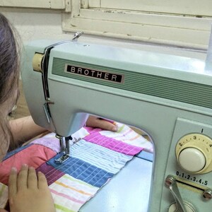 May include: A person is using a blue Brother sewing machine to sew a colorful patchwork quilt.