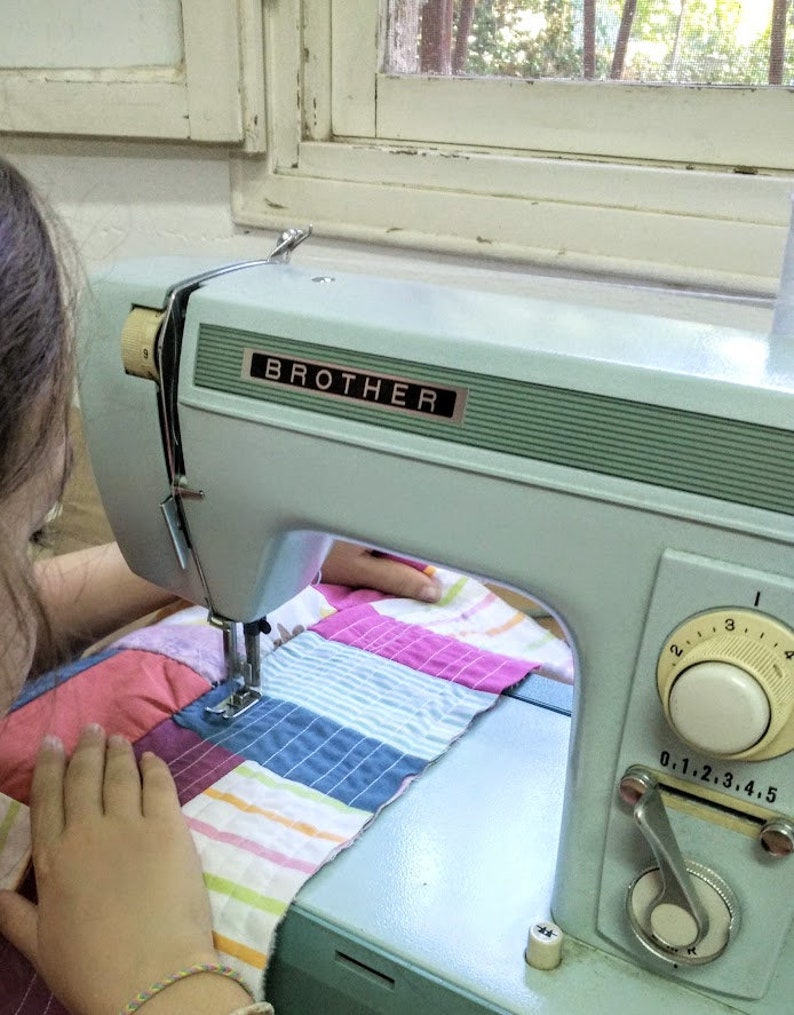 May include: A person is using a blue Brother sewing machine to sew a colorful patchwork quilt.