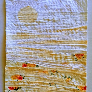 Pu&ograve; includere: Un arazzo da parete in tessuto bianco con un design cucito di un sole giallo e un paesaggio con fiori gialli e arancioni. Il tessuto ha un aspetto testurizzato e stratificato.