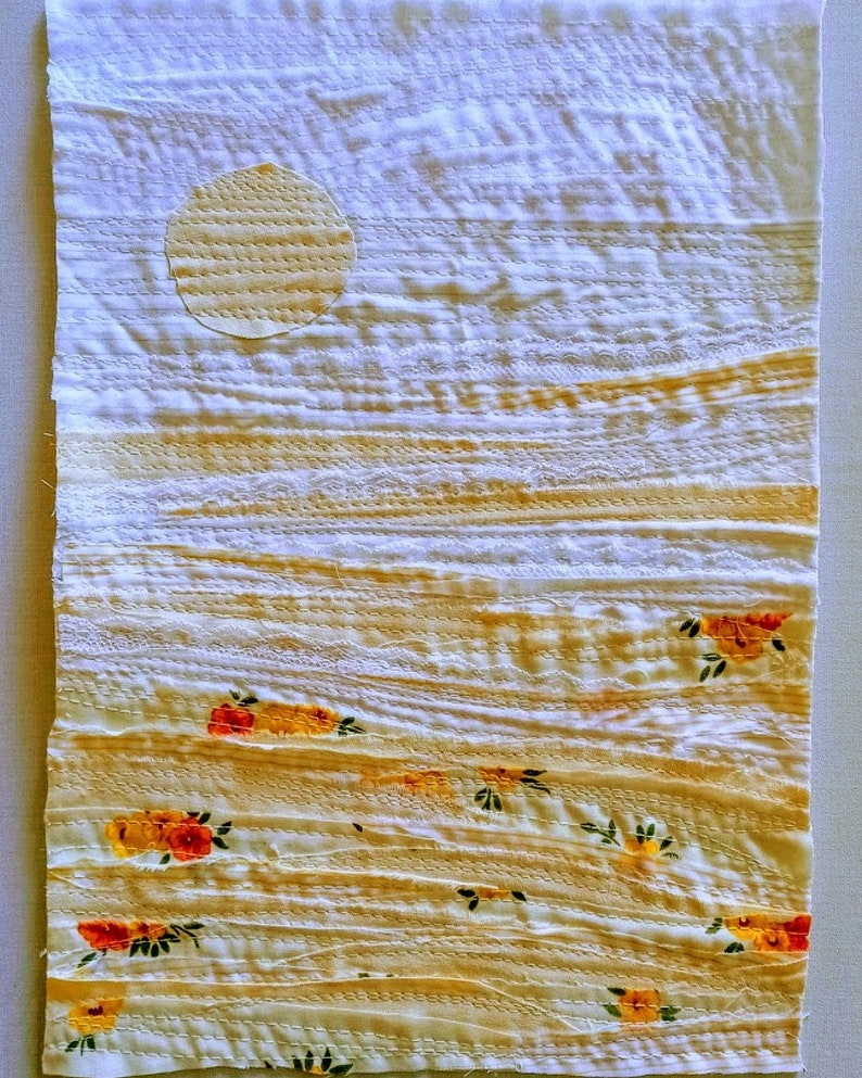 Pu&ograve; includere: Un arazzo da parete in tessuto bianco con un design cucito di un sole giallo e un paesaggio con fiori gialli e arancioni. Il tessuto ha un aspetto testurizzato e stratificato.