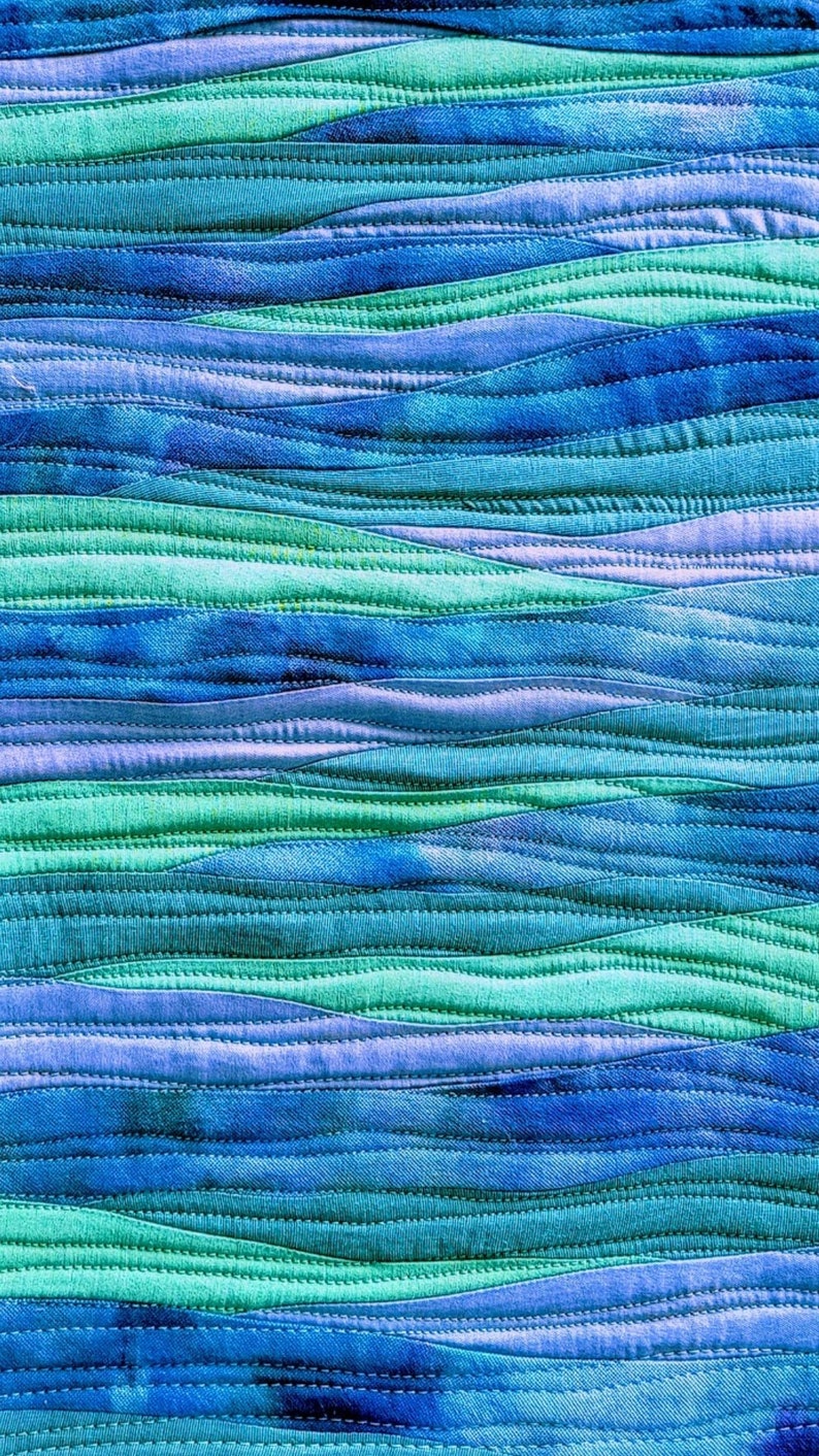Op de afbeelding: Een close-up van een blauw en groen stoffen quilt met een golvend patroon. De stof is in stroken aan elkaar genaaid en de naden zijn zichtbaar. De quilt is gemaakt van een zacht, getextureerd materiaal.