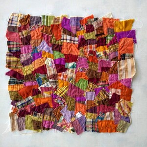 May include: A patchwork textile composed of various fabric scraps in shades of orange, purple, green, and red. The irregular shapes and textures create a vibrant, handcrafted appearance. The textile is likely a small quilt or decorative piece.