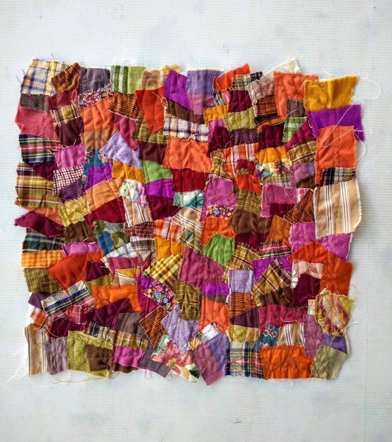 May include: A patchwork textile composed of various fabric scraps in shades of orange, purple, green, and red. The irregular shapes and textures create a vibrant, handcrafted appearance. The textile is likely a small quilt or decorative piece.