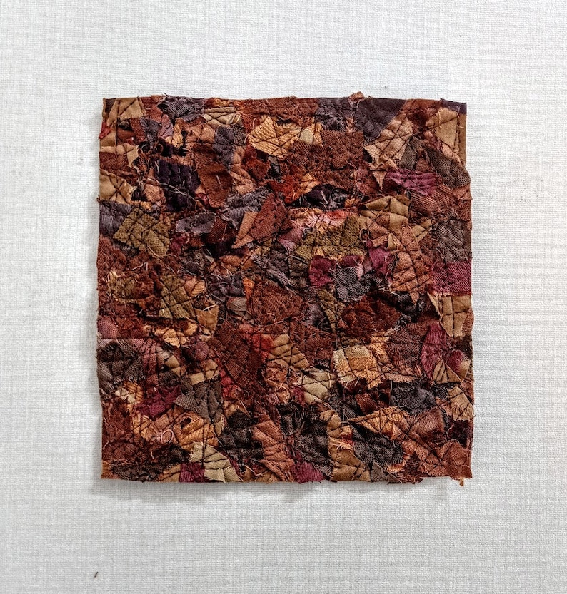 May include: A square piece of fabric art made from many small pieces of brown, red, and green fabric sewn together.