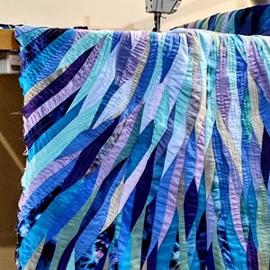 Op de afbeelding: Een close-up van een naaimachine die een blauwe en paarse quilt naait. De quilt is gemaakt van stroken stof die in een golvend patroon aan elkaar zijn genaaid.