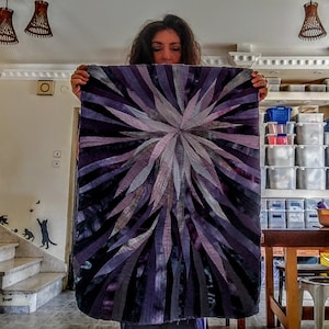 Op de afbeelding: Een groot, abstract quilt met een stervormig patroon in tinten paars, grijs en wit. De quilt is gemaakt van stofstroken die aan elkaar zijn genaaid om een ​​getextureerd effect te cre&euml;ren.
