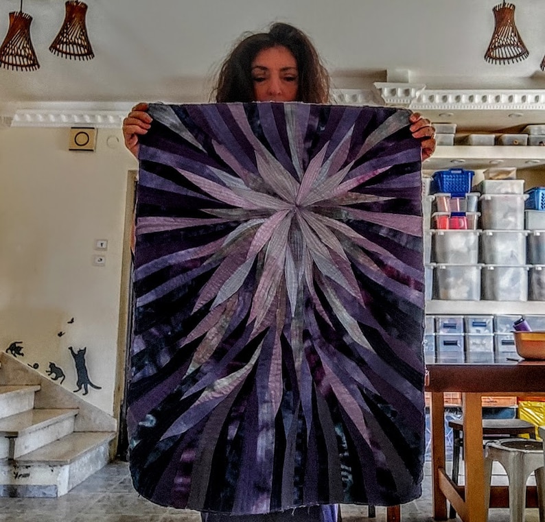 Op de afbeelding: Een groot, abstract quilt met een stervormig patroon in tinten paars, grijs en wit. De quilt is gemaakt van stofstroken die aan elkaar zijn genaaid om een ​​getextureerd effect te cre&euml;ren.