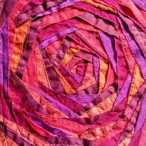 Pu&ograve; includere: Primo piano di un'opera d'arte tessile circolare realizzata con strisce di tessuto in tonalit&agrave; rosa, viola e arancio. Il tessuto &egrave; cucito a spirale, creando un design testurizzato e colorato.