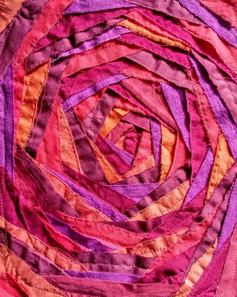 Pu&ograve; includere: Primo piano di un'opera d'arte tessile circolare realizzata con strisce di tessuto in tonalit&agrave; rosa, viola e arancio. Il tessuto &egrave; cucito a spirale, creando un design testurizzato e colorato.