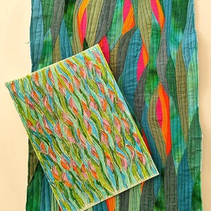 May include: A close-up of a hand-stitched fabric art piece with a green, blue, orange, and pink colour scheme. The fabric is arranged in a wavy pattern, resembling leaves or water. A smaller drawing of a similar design is placed on top of the fabric.