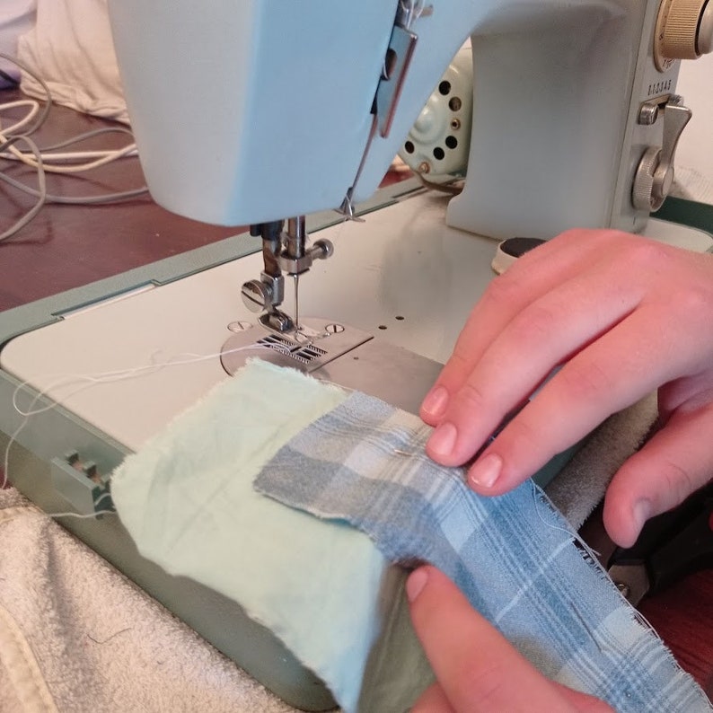 May include: A close-up of a sewing machine with a light blue body and a white thread. A person is holding a piece of fabric with a blue and white plaid pattern.