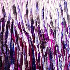 May include: A close-up of a fabric art piece with a textured surface. The fabric is made up of many strips of purple, blue, and white fabric that are sewn together. The fabric is arranged in a vertical pattern, creating a sense of depth and movement.