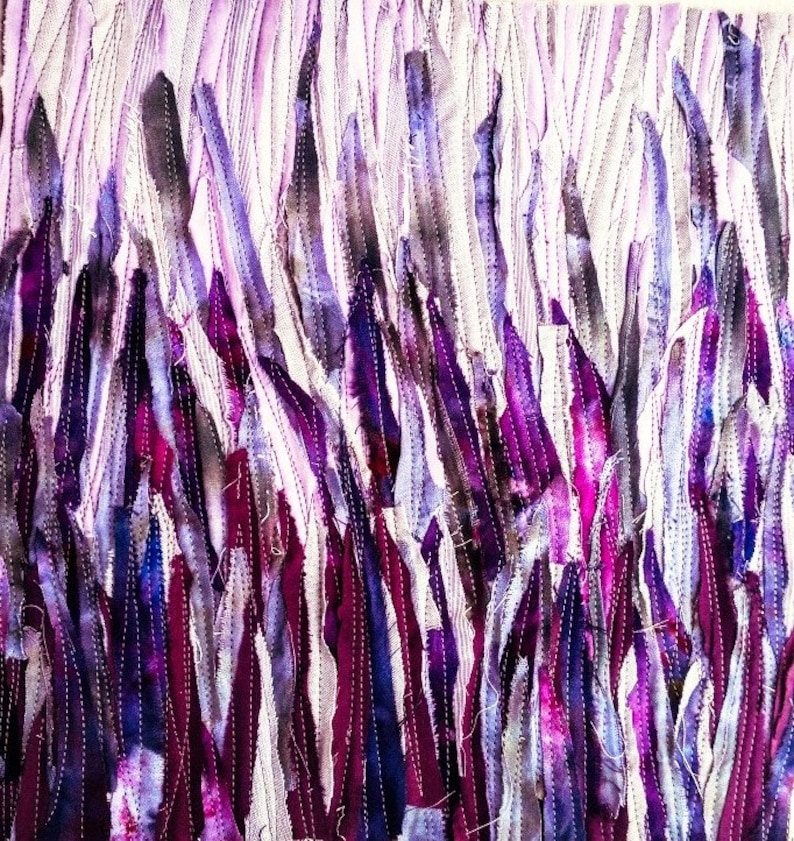 May include: A close-up of a fabric art piece with a textured surface. The fabric is made up of many strips of purple, blue, and white fabric that are sewn together. The fabric is arranged in a vertical pattern, creating a sense of depth and movement.