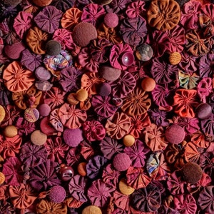 Pu&ograve; includere: Primo piano di una collezione di fiori di stoffa in tonalit&agrave; di rosso, rosa e marrone. I fiori sono disposti in un motivo casuale e creano uno sfondo testurizzato e colorato.