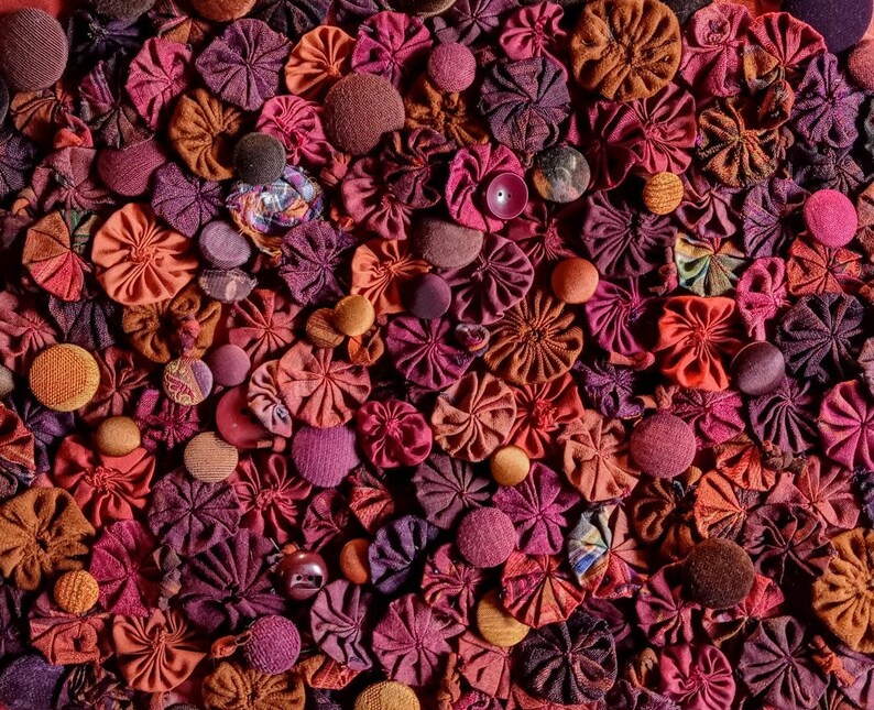 Pu&ograve; includere: Primo piano di una collezione di fiori di stoffa in tonalit&agrave; di rosso, rosa e marrone. I fiori sono disposti in un motivo casuale e creano uno sfondo testurizzato e colorato.