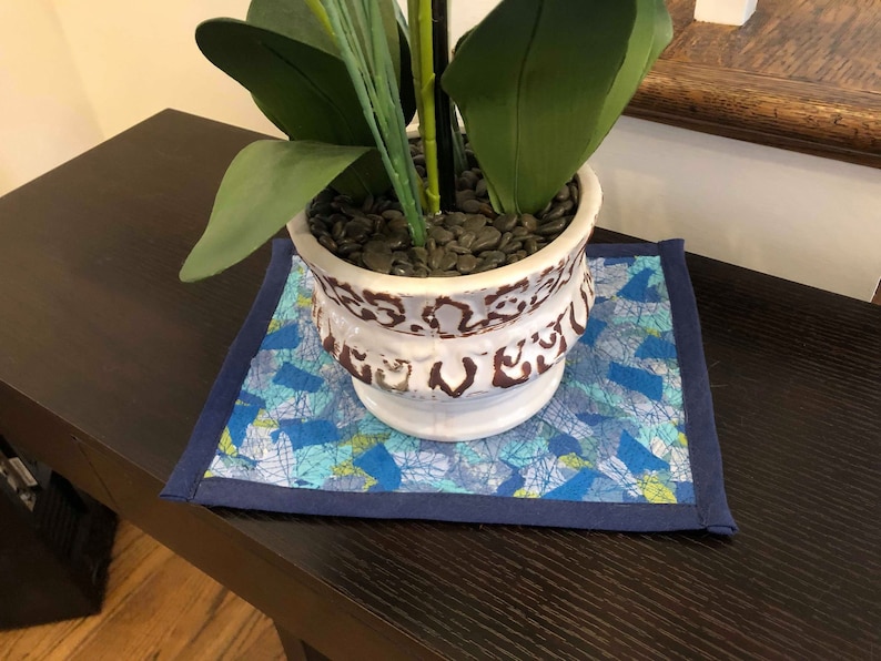 May include: A square, quilted plant mat with a blue and white abstract pattern and a navy blue border. A white and brown ceramic pot with a plant sits on the mat. The pot is filled with black pebbles.