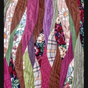 May include: A quilt made from assorted fabric scraps in shades of brown, purple, green, and red. The fabric is sewn together in a wavy pattern, creating a unique and textured design.