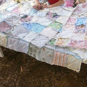 May include: A patchwork quilt made from a variety of fabrics in shades of pink, blue, and white. The quilt is made up of squares and rectangles, and the edges are frayed. The quilt is lying on a table.