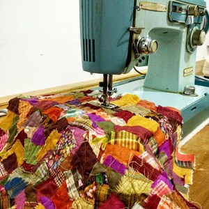 May include: A vintage turquoise sewing machine stitching a colorful patchwork quilt. The quilt is composed of various fabric scraps in shades of orange, purple, yellow, and green. The machine is set on a wooden surface.
