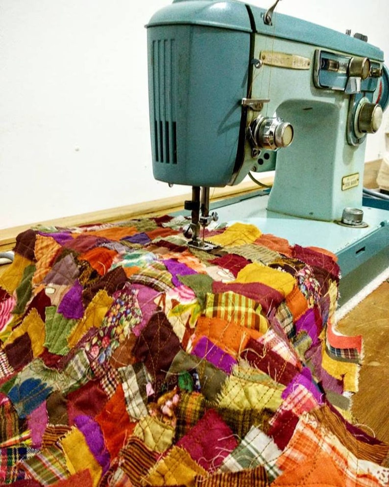 May include: A vintage turquoise sewing machine stitching a colorful patchwork quilt. The quilt is composed of various fabric scraps in shades of orange, purple, yellow, and green. The machine is set on a wooden surface.