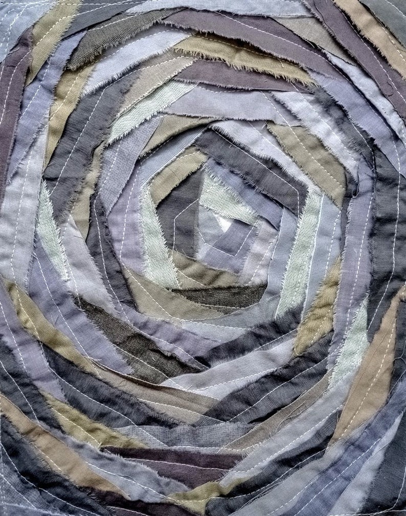 Pu&ograve; includere: Un'opera d'arte tessile circolare realizzata con strisce di tessuto in tonalit&agrave; di grigio, marrone e beige. Le strisce di tessuto sono cucite insieme a spirale, creando un design visivamente interessante e testurizzato.
