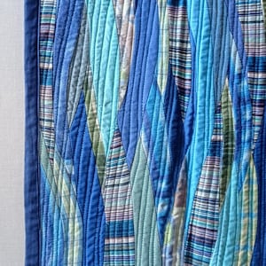 Op de afbeelding: Een blauwe en groene gequilte wandhanger met een golvend patroon. De stof is gemaakt van verschillende tinten blauw en groen, en de quilt is in een golvend patroon gemaakt. De wandhanger is omlijst met een donkerblauwe rand.