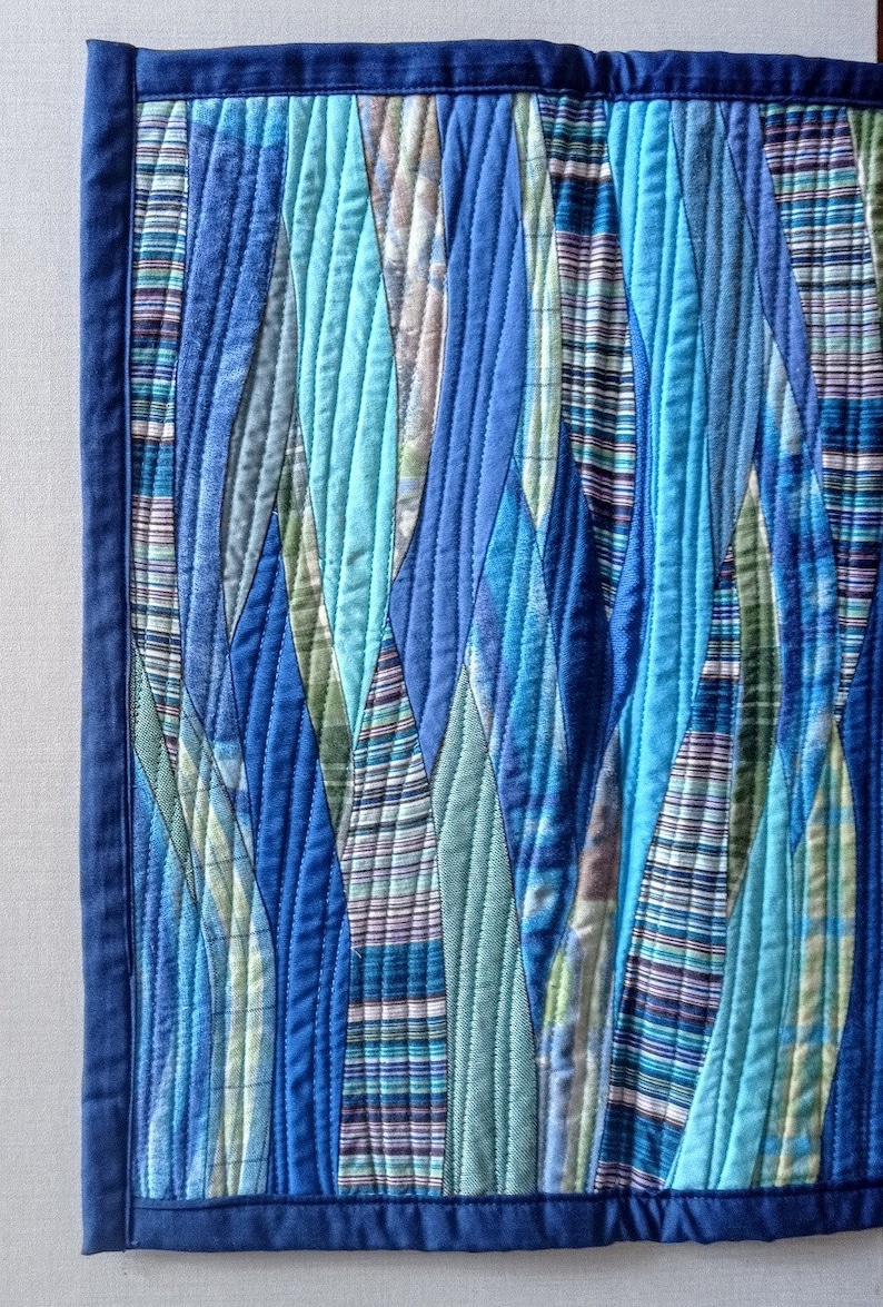 Op de afbeelding: Een blauwe en groene gequilte wandhanger met een golvend patroon. De stof is gemaakt van verschillende tinten blauw en groen, en de quilt is in een golvend patroon gemaakt. De wandhanger is omlijst met een donkerblauwe rand.