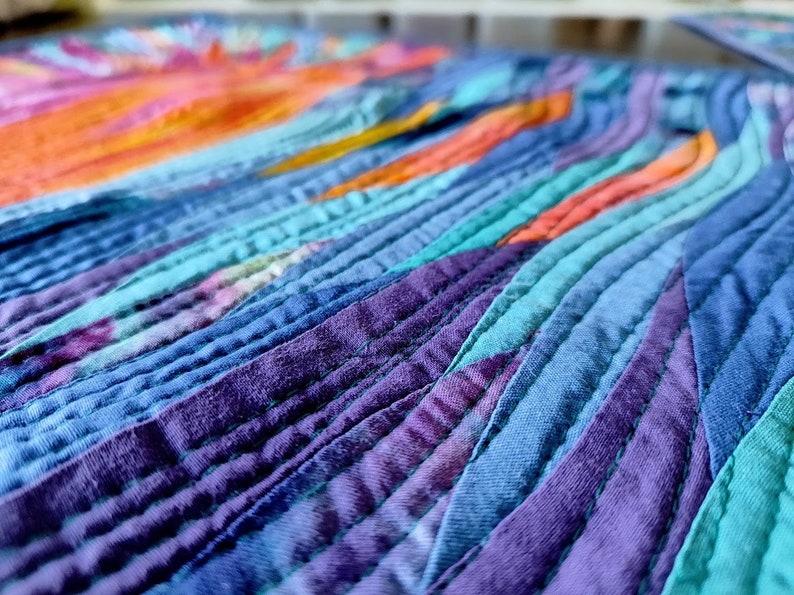 May include: Close-up of a colourful fabric quilt with a wavy pattern. The quilt is made of strips of fabric in shades of blue, purple, teal, orange, and pink. The fabric is sewn together in a way that creates a flowing, organic design.
