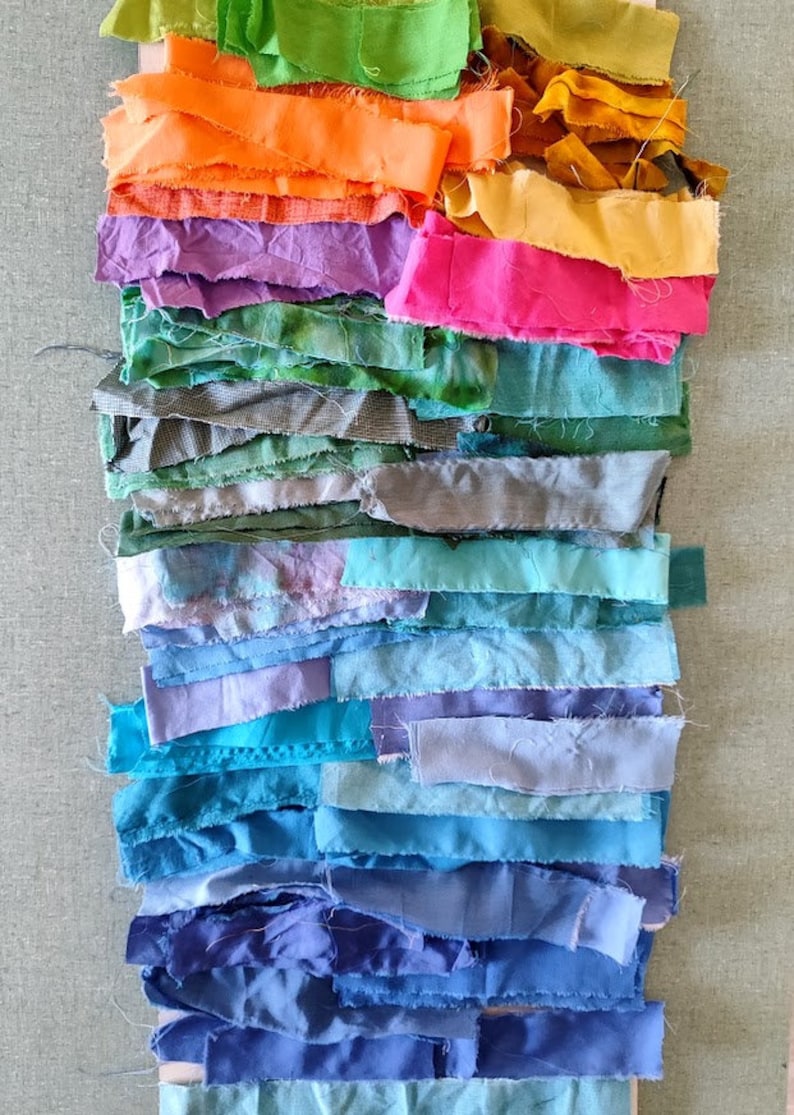 May include: A collection of fabric scraps in various shades of green, orange, purple, pink, blue, and teal. The fabric scraps are arranged in a rainbow pattern, with the darkest colours at the bottom and the lightest colours at the top.