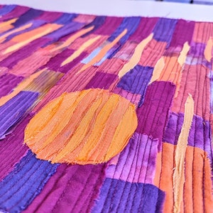 May include: A close-up of a fabric art piece with a patchwork design. The piece is made up of strips of fabric in shades of purple, pink, and orange. A large orange circle is in the centre of the piece.