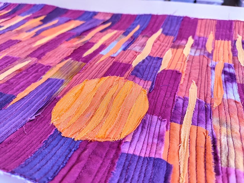 May include: A close-up of a fabric art piece with a patchwork design. The piece is made up of strips of fabric in shades of purple, pink, and orange. A large orange circle is in the centre of the piece.