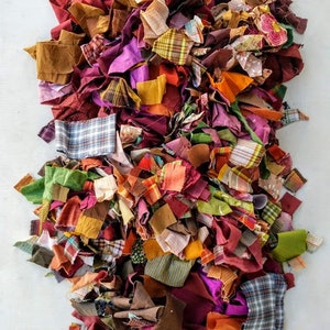 Op de afbeelding: Een grote stapel kleurrijke stoffenresten in verschillende tinten rood, oranje, geel, groen, bruin en paars. De stoffenresten zijn in verschillende patronen en texturen, waaronder ruit, gestreept en effen.