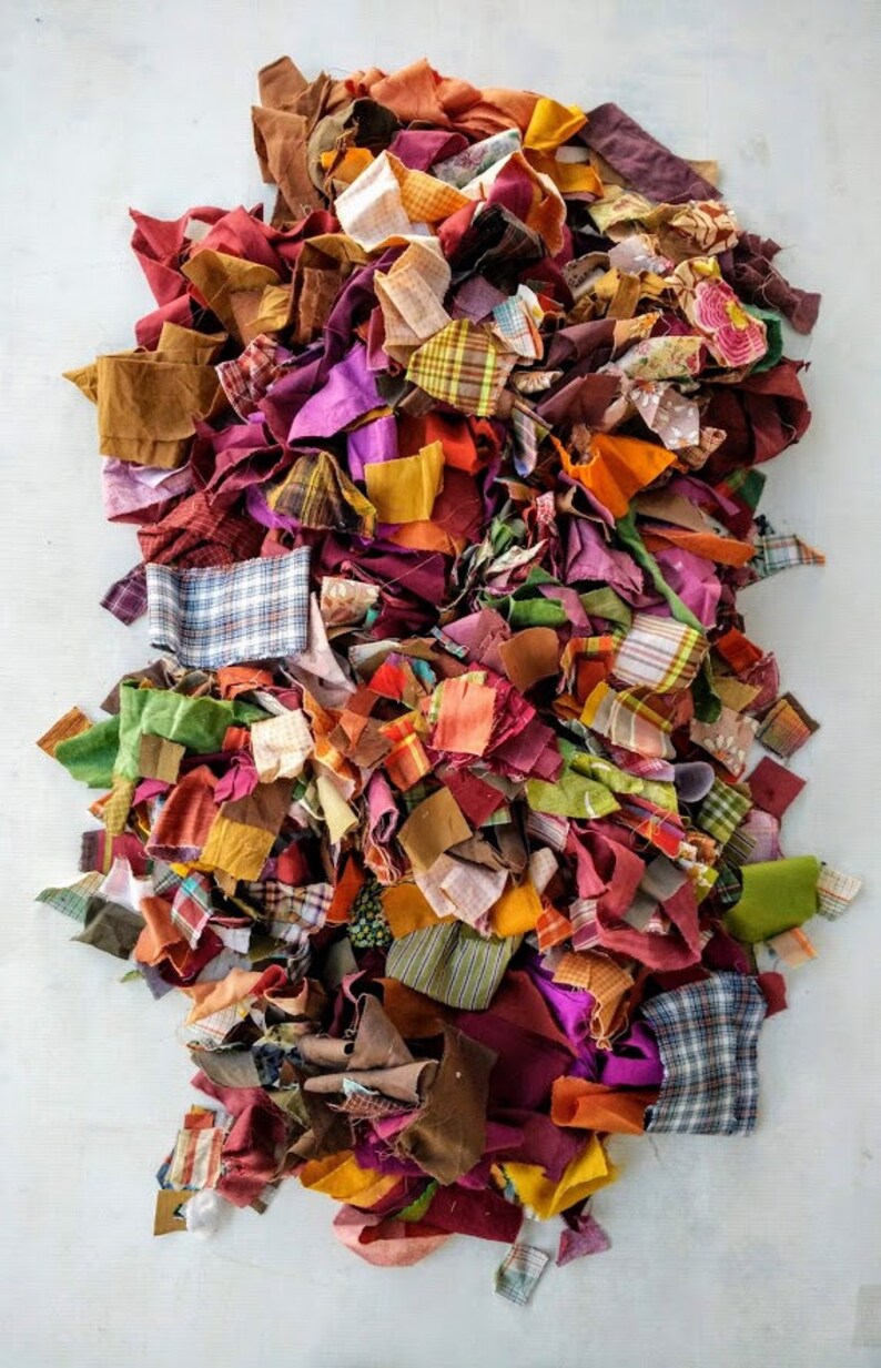 Op de afbeelding: Een grote stapel kleurrijke stoffenresten in verschillende tinten rood, oranje, geel, groen, bruin en paars. De stoffenresten zijn in verschillende patronen en texturen, waaronder ruit, gestreept en effen.