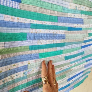 Puede incluir: Una colcha hecha de tiras de tela en tonos de azul, verde y blanco. La tela est&aacute; cosida en un patr&oacute;n horizontal, creando un efecto a rayas. La colcha est&aacute; hecha con una variedad de telas diferentes, incluyendo estampados florales, rayas y lisos.