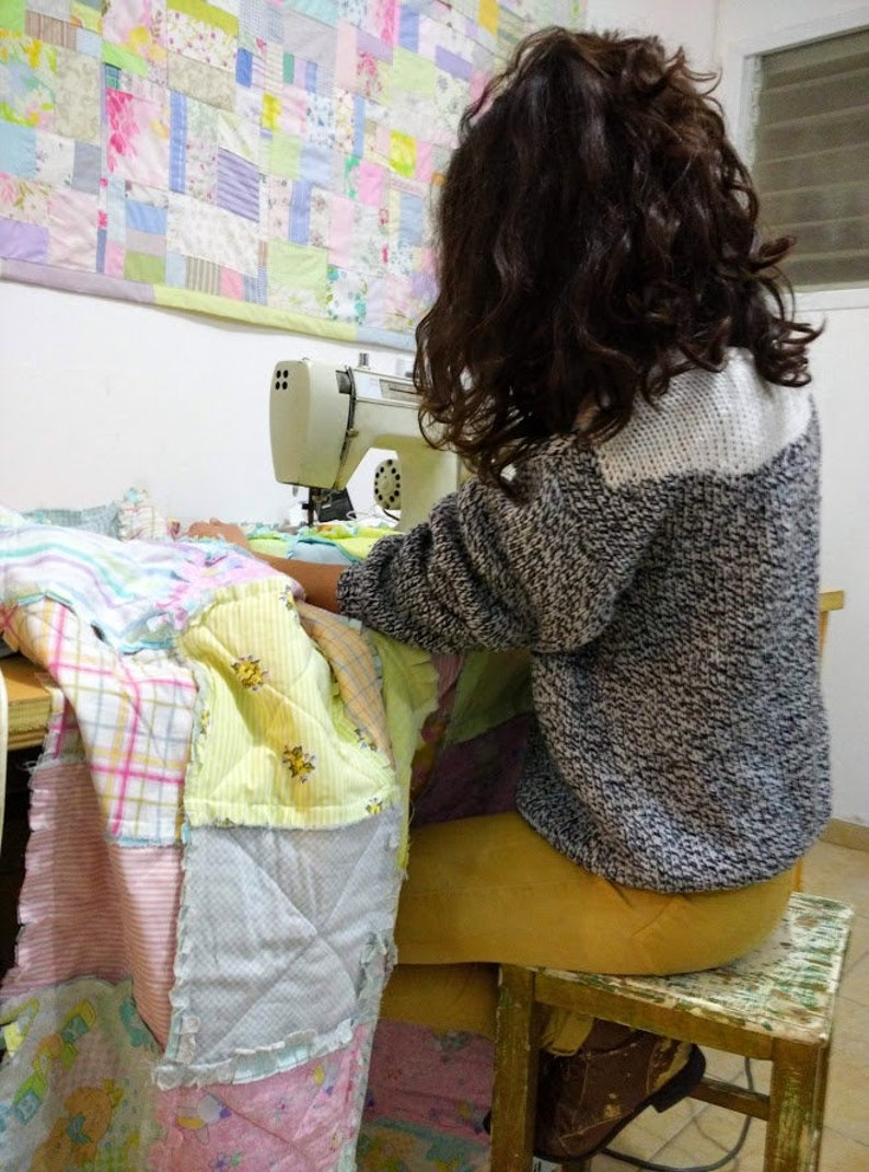 May include: A person is sitting on a stool and using a white sewing machine to sew a patchwork quilt made of various colours and patterns.