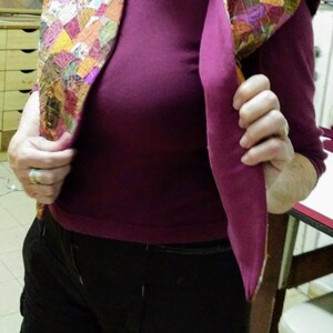 Op de afbeelding: Een vrouw draagt een paarse blouse en een zwarte broek. Ze draagt een kleurrijk patchwork vest met een capuchon.