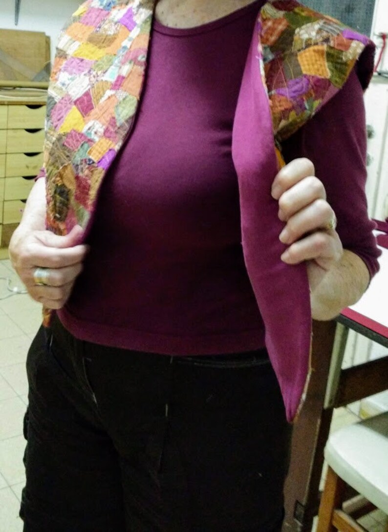 Op de afbeelding: Een vrouw draagt een paarse blouse en een zwarte broek. Ze draagt een kleurrijk patchwork vest met een capuchon.