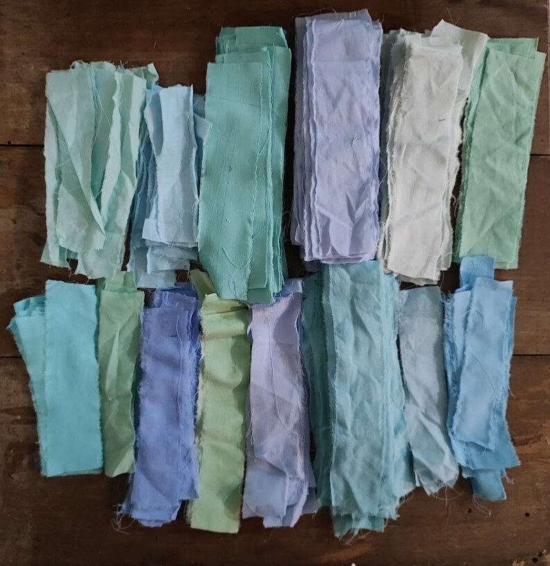 Puede incluir: Una colecci&oacute;n de recortes de tela en varios tonos de azul y verde. La tela est&aacute; cortada en tiras largas y delgadas y dispuestas en filas.