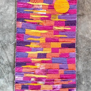 May include: A colourful patchwork rug with a yellow sun design. The rug is made of different shades of pink, purple, and orange fabric.