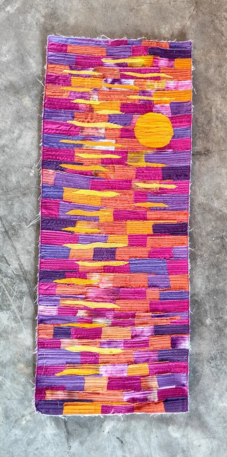May include: A colourful patchwork rug with a yellow sun design. The rug is made of different shades of pink, purple, and orange fabric.