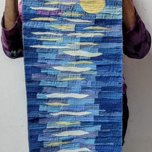 May include: A long, rectangular wall hanging made of fabric with a blue, white and yellow abstract design. The design features a large yellow circle in the centre, surrounded by white and blue lines.