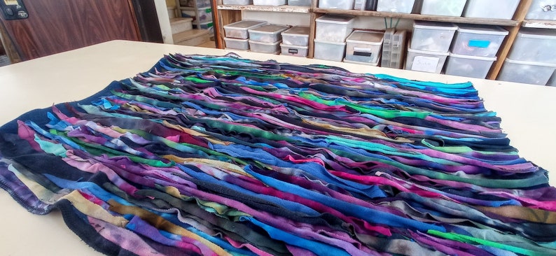 Puede incluir: Una alfombra colorida tejida a mano con tiras de tela en tonos azules, morados, verdes y marrones. La alfombra est&aacute; extendida sobre una superficie blanca.