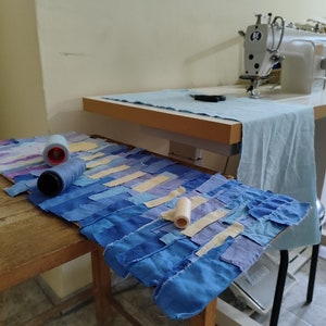 May include: A close-up of a patchwork quilt made of blue, purple, and cream fabric scraps. The quilt is laid out on a wooden table with spools of thread and a sewing machine in the background.