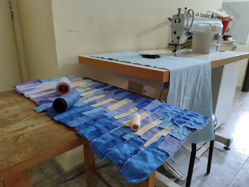 May include: A close-up of a patchwork quilt made of blue, purple, and cream fabric scraps. The quilt is laid out on a wooden table with spools of thread and a sewing machine in the background.