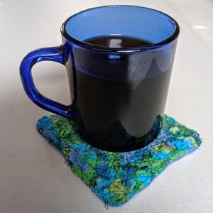 May include: A blue ceramic mug with a handle filled with dark coffee or tea, sitting on a colourful patchwork fabric coaster.