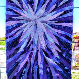 Op de afbeelding: Een paarse en blauwe quilt met een stervormig ontwerp. De quilt is gemaakt van veel verschillende stoffen en is gestikt met een fijne steek.
