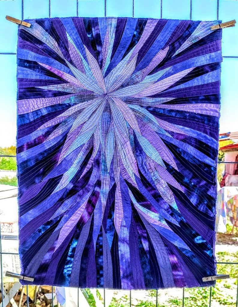 Op de afbeelding: Een paarse en blauwe quilt met een stervormig ontwerp. De quilt is gemaakt van veel verschillende stoffen en is gestikt met een fijne steek.