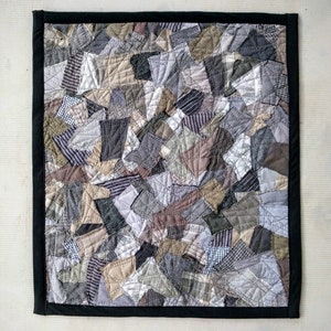 Może przedstawiać: Ręcznie robiona patchworkowa kołdra z czarną obw&oacute;dką. Kołdra składa się z r&oacute;żnych skrawk&oacute;w materiału w odcieniach szarości, brązu i beżu, tworząc teksturowany, abstrakcyjny wz&oacute;r. Kołdra jest prawdopodobnie używana do dekoracji wnętrz.