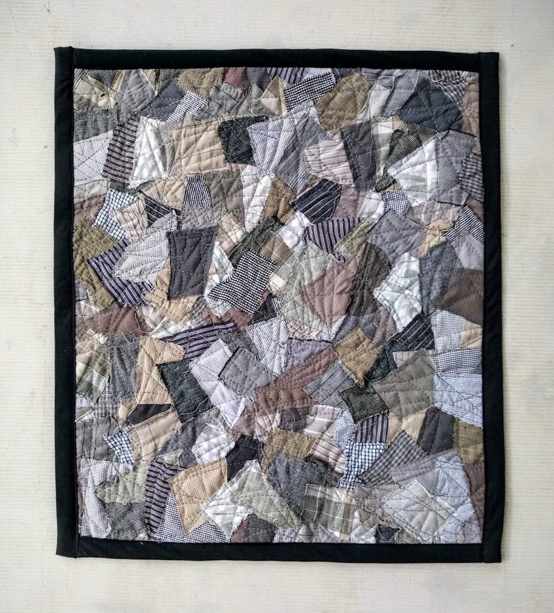 Może przedstawiać: Ręcznie robiona patchworkowa kołdra z czarną obw&oacute;dką. Kołdra składa się z r&oacute;żnych skrawk&oacute;w materiału w odcieniach szarości, brązu i beżu, tworząc teksturowany, abstrakcyjny wz&oacute;r. Kołdra jest prawdopodobnie używana do dekoracji wnętrz.