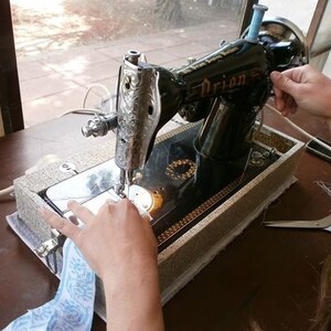 May include: A black sewing machine with a silver design and the word "Orion" on the side. The machine is on a wooden table with a white cloth and a pair of scissors.